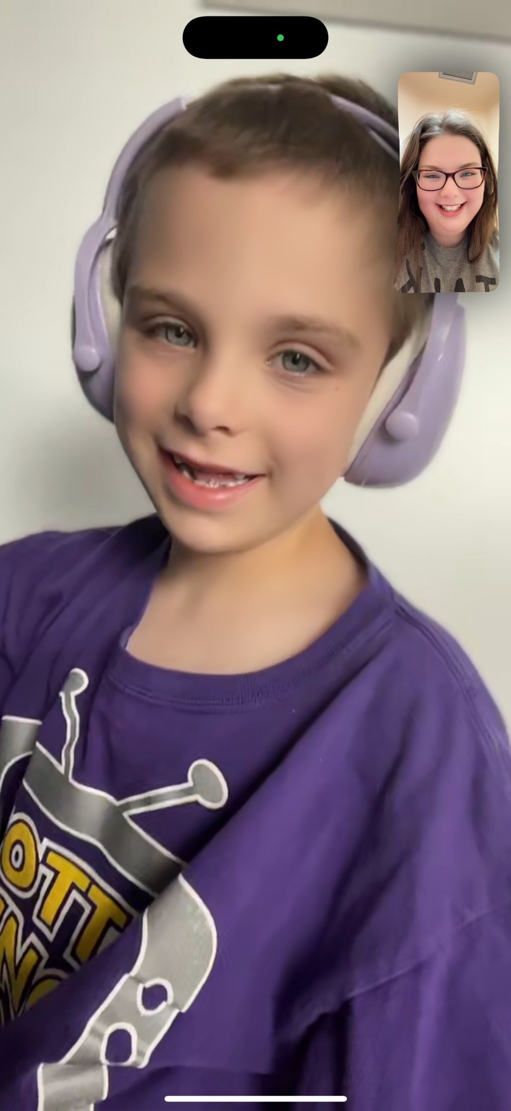 A smiling young boy wearing purple headphones and a purple shirt during a video call with his mom, who appears in the corner of the screen smiling back.
