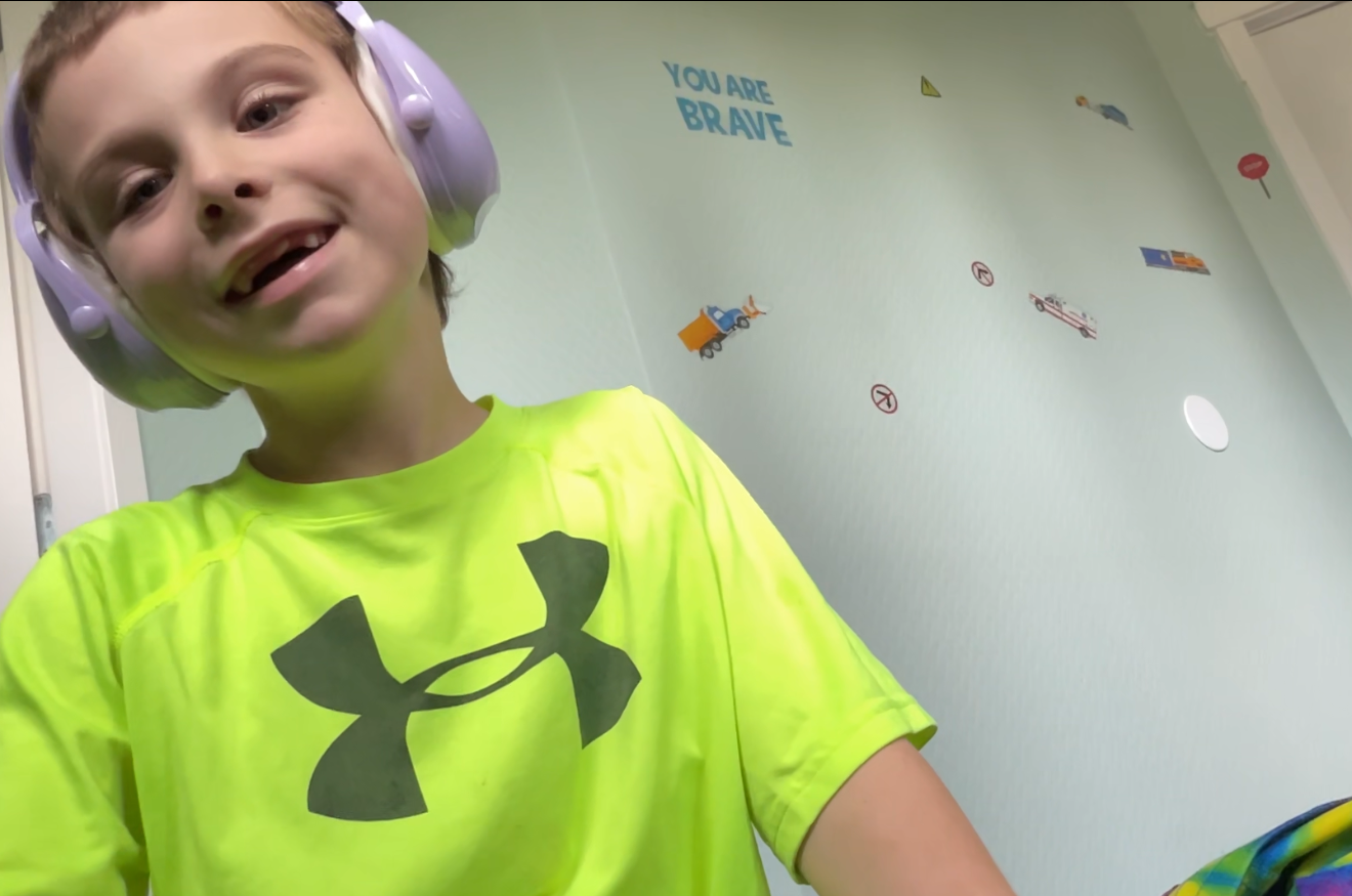 Young boy wearing headphones and a bright green shirt smiling during a FaceTime call, with wall decals behind him that say “You are brave.”